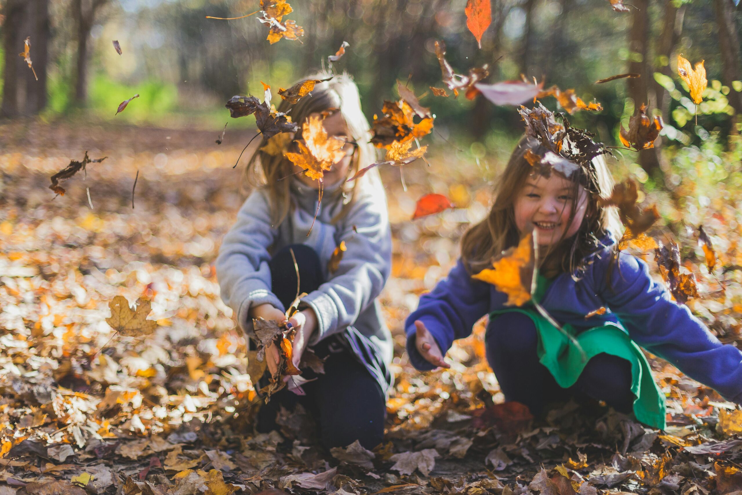 Two smiling girls playing joyfully amidst colorful fall leaves in a sunlit forest