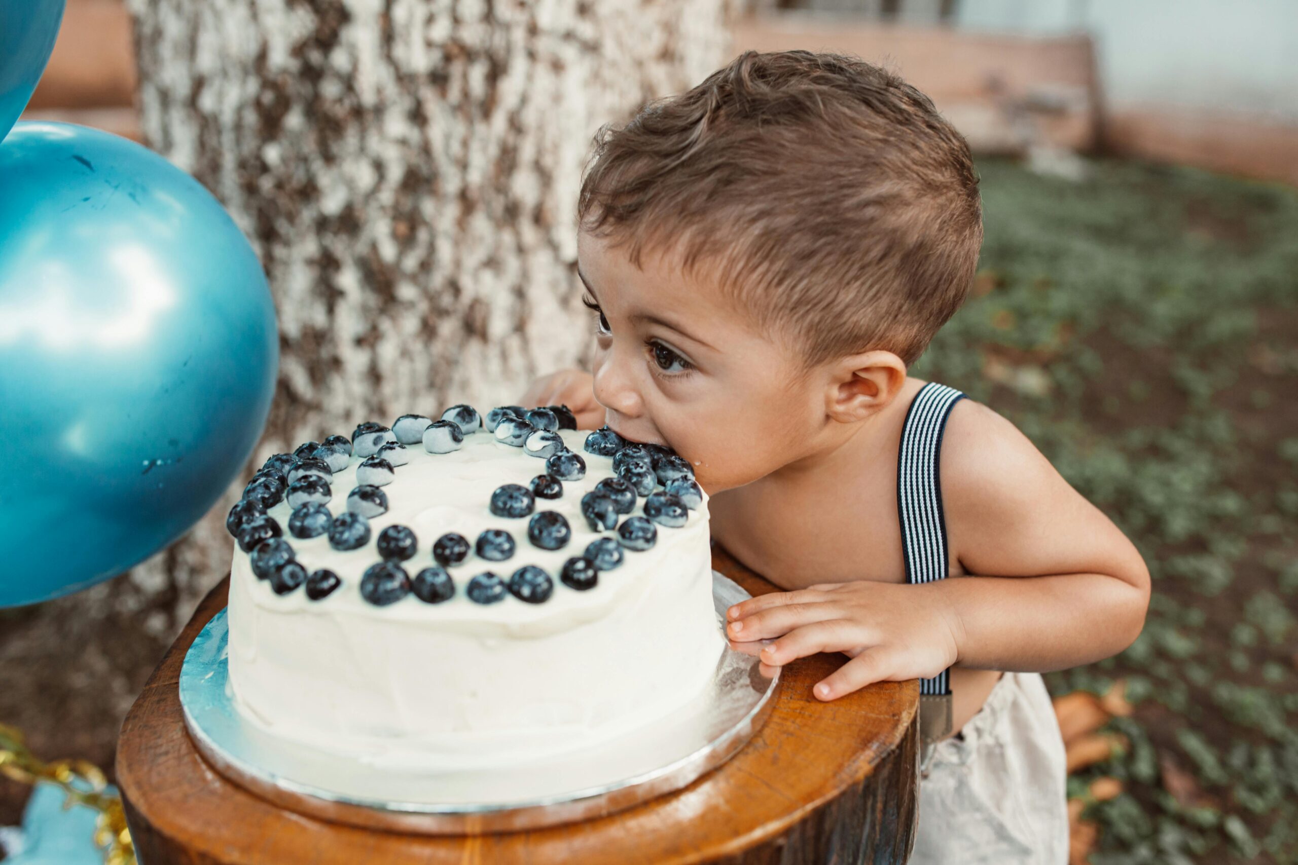 A young child delightfully devours a blueberry-topped cake outdoors, celebrating a special occasion.