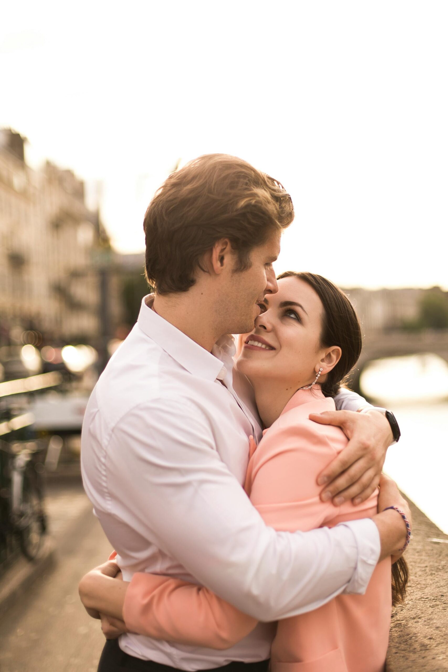 A loving couple embraces by a riverside as the sun sets, capturing a moment of affection.