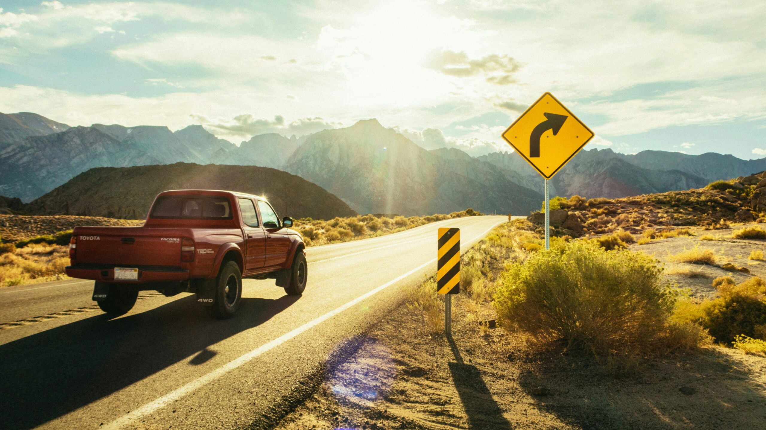 A red pickup truck driving on a sunlit road with mountains in the background and a road sign ahead.