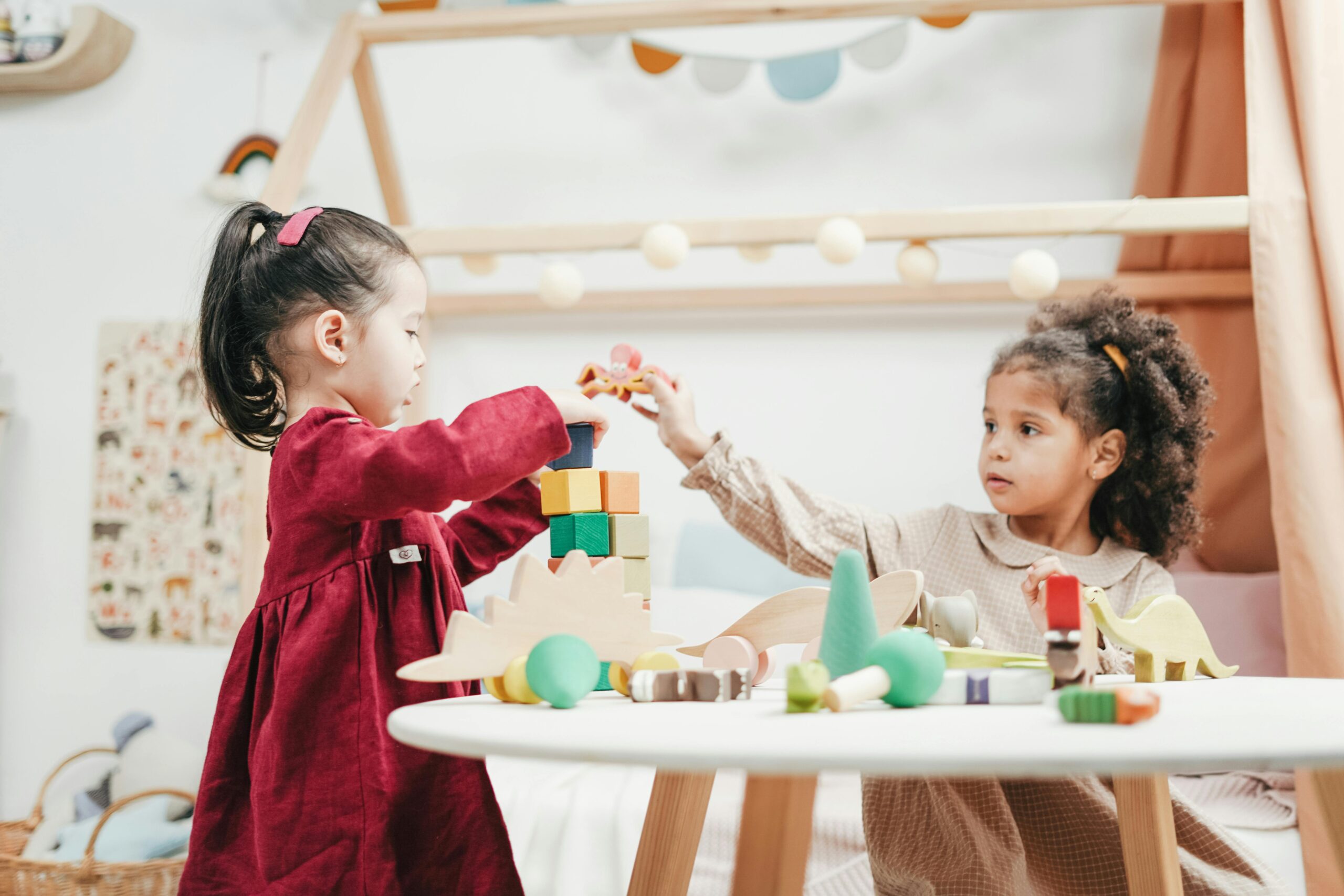 Two young girls enjoying playtime with wooden toys indoors in a warm, colorful playroom.