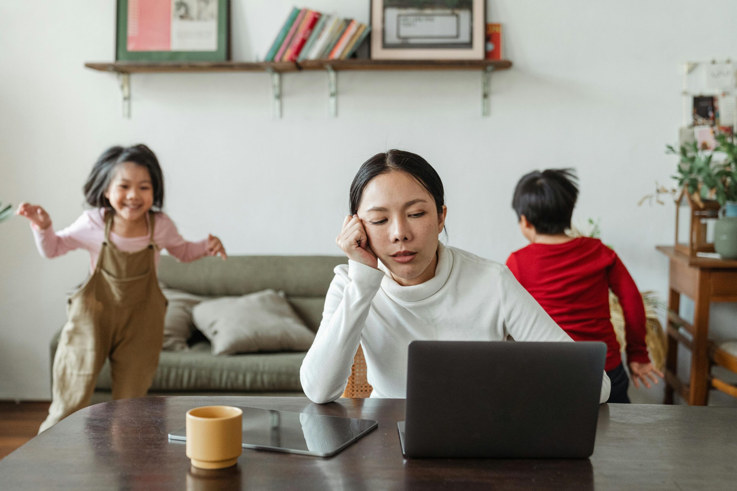 A tired mother working on a laptop while her children play around indoors, highlighting remote work challenges.