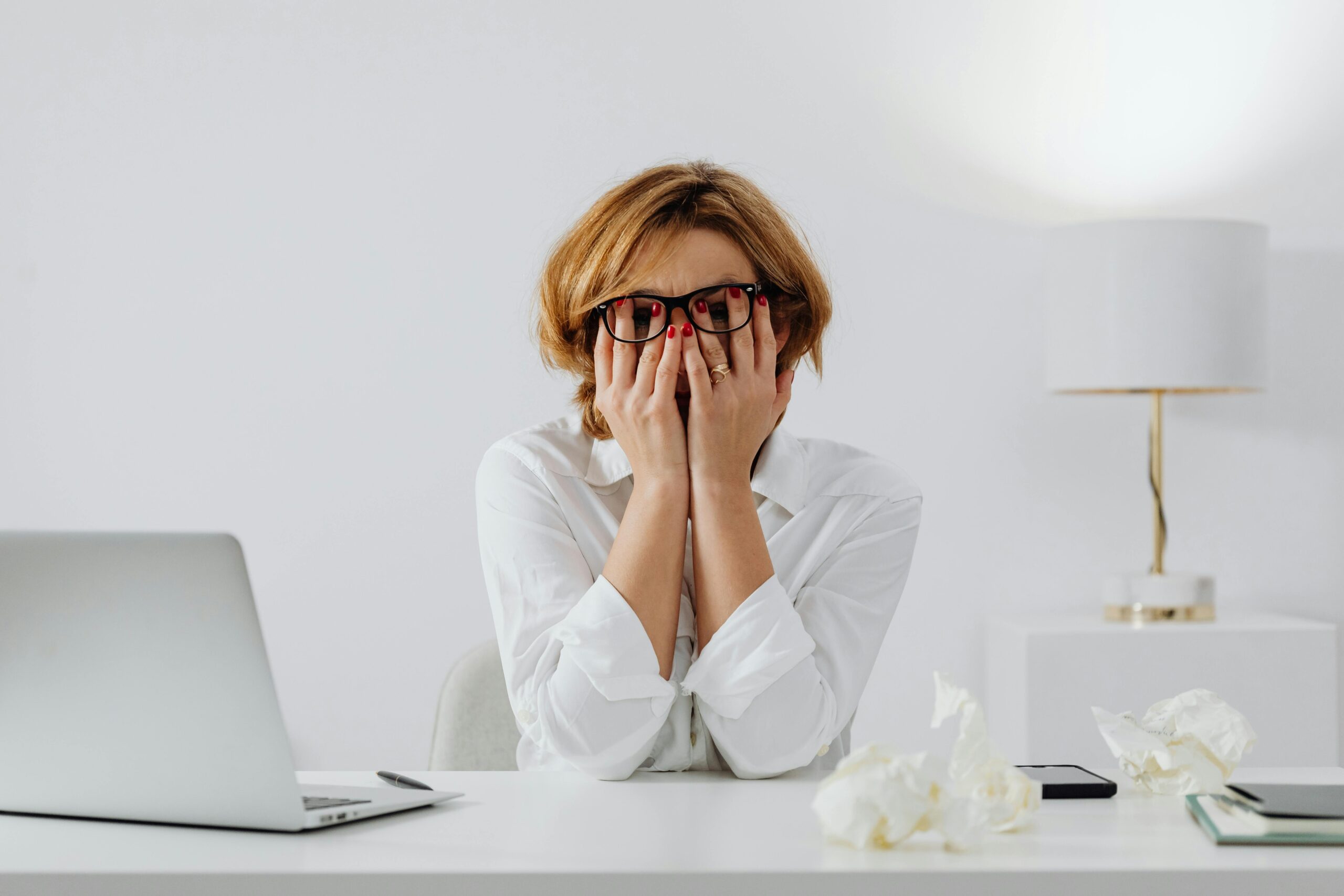 Woman with hands on face, sitting at desk with laptop and tissues, feeling stressed.