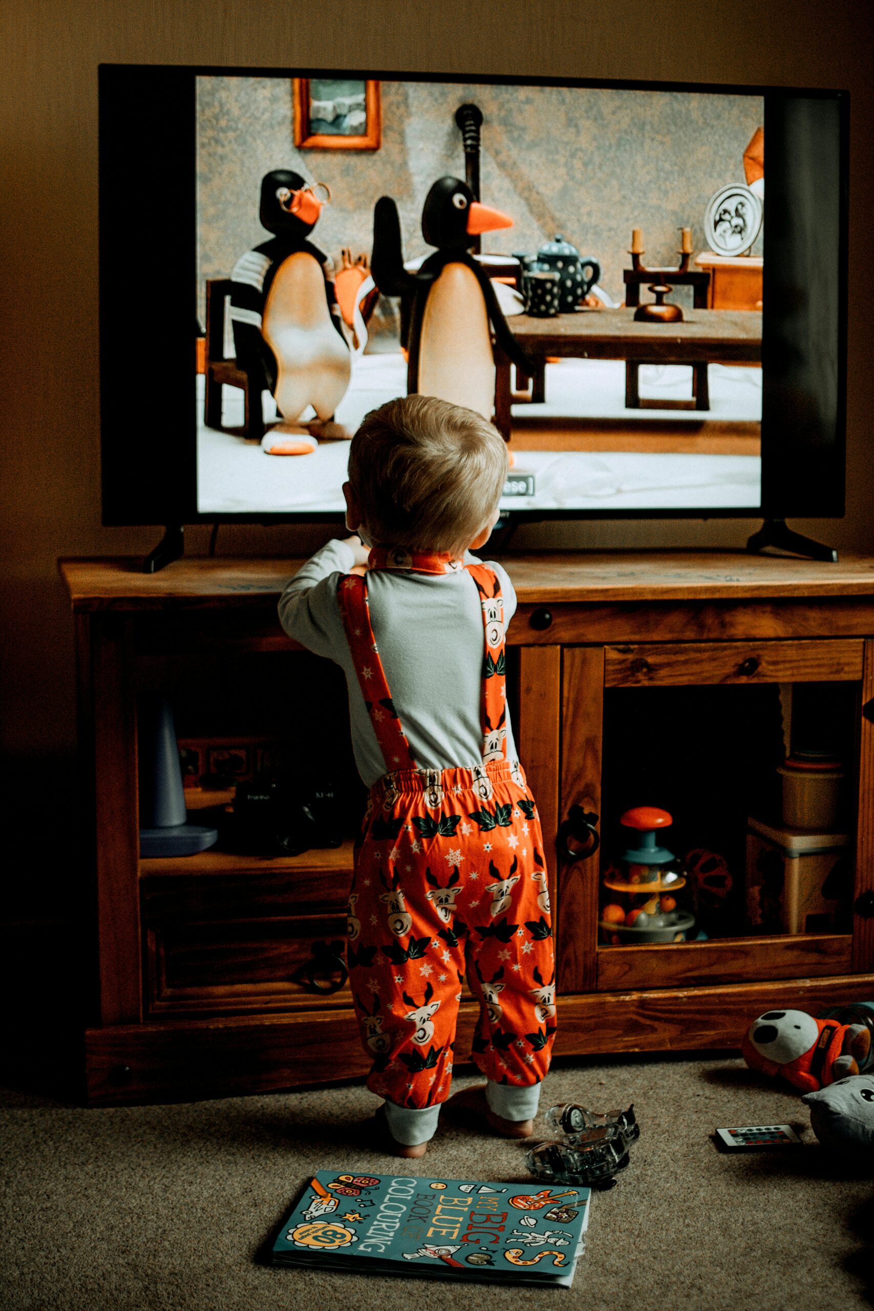 A toddler in colorful pajamas watches an animated penguin show on television indoors.