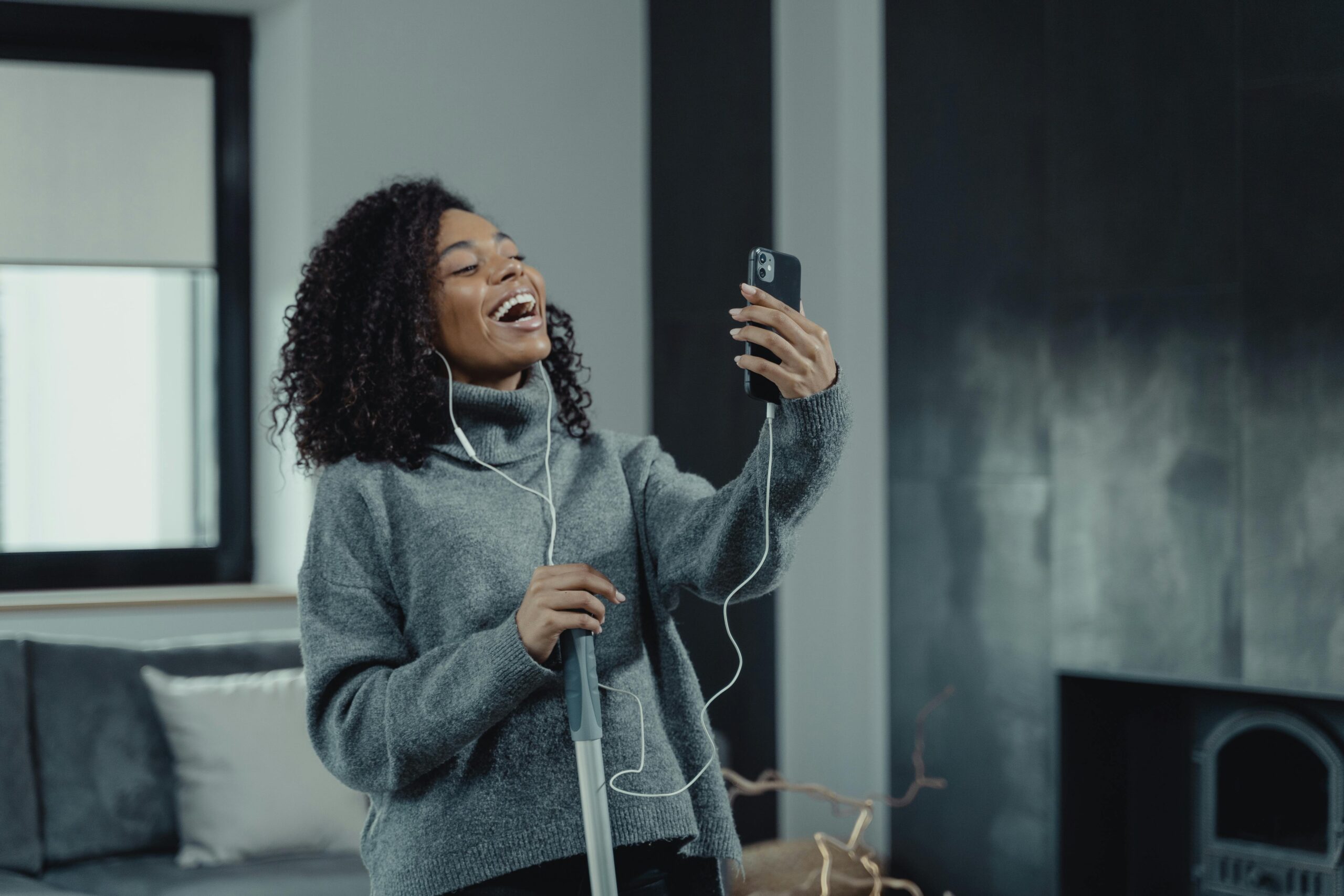 Happy woman with curly hair using smartphone for video call while cleaning her house.