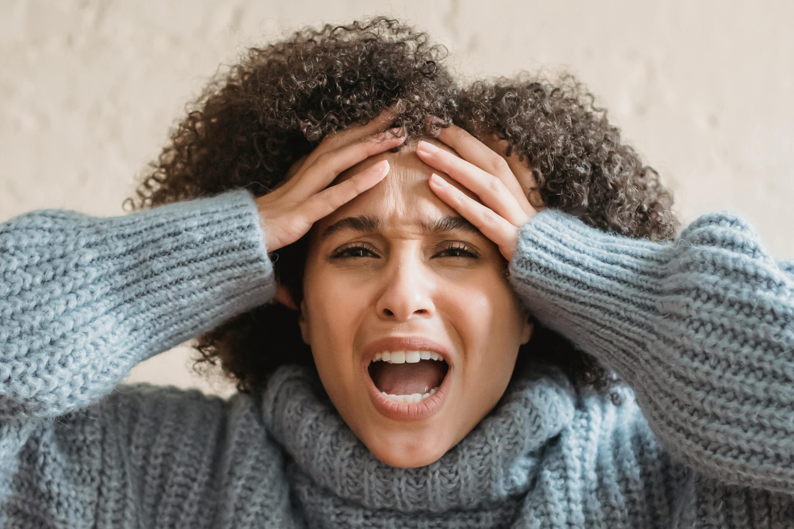 Expressive African American woman with curly brown hair touching head and yelling against light background