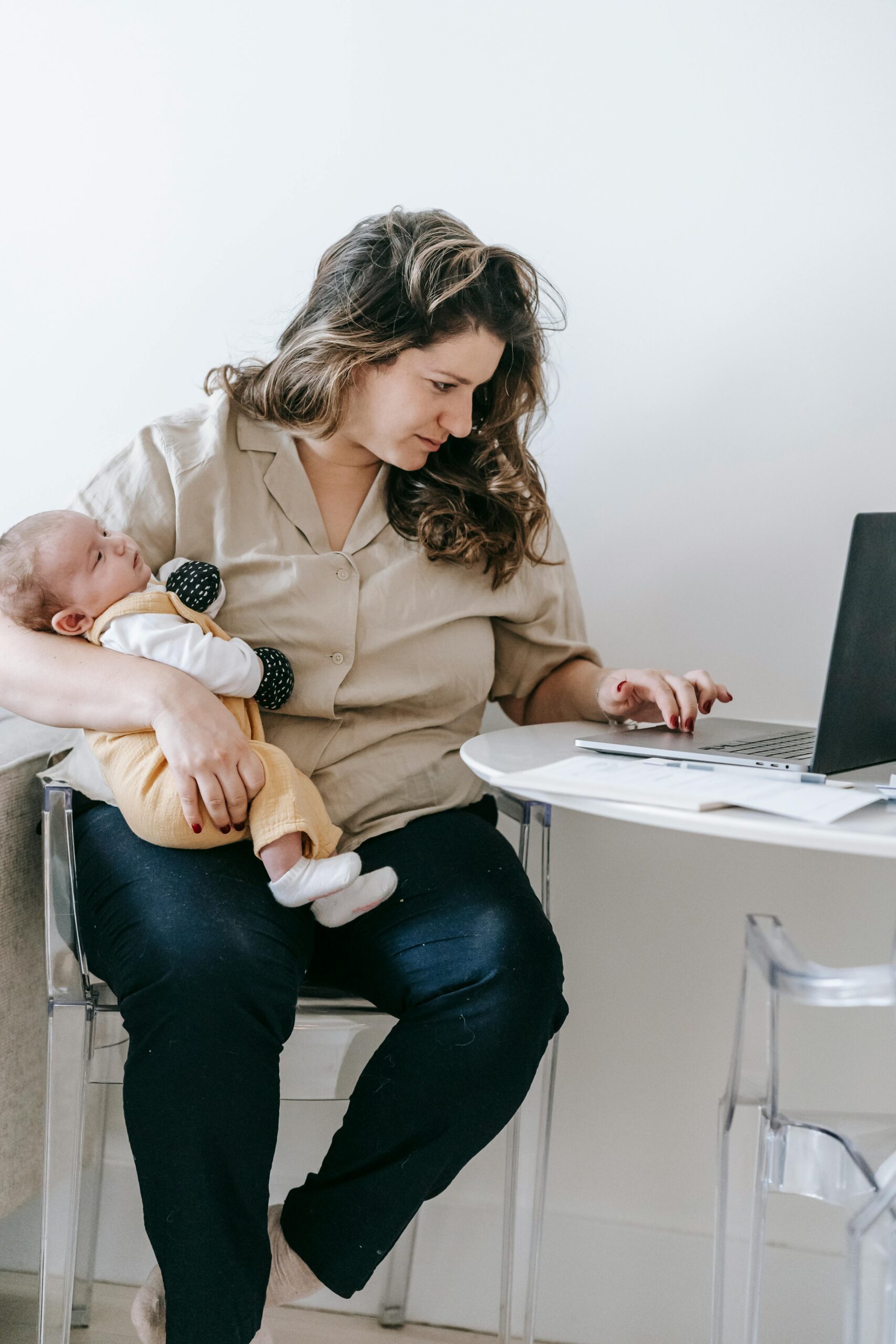 Concentrated mom sitting on chair with cute baby and browsing laptop while working remotely on maternity leave