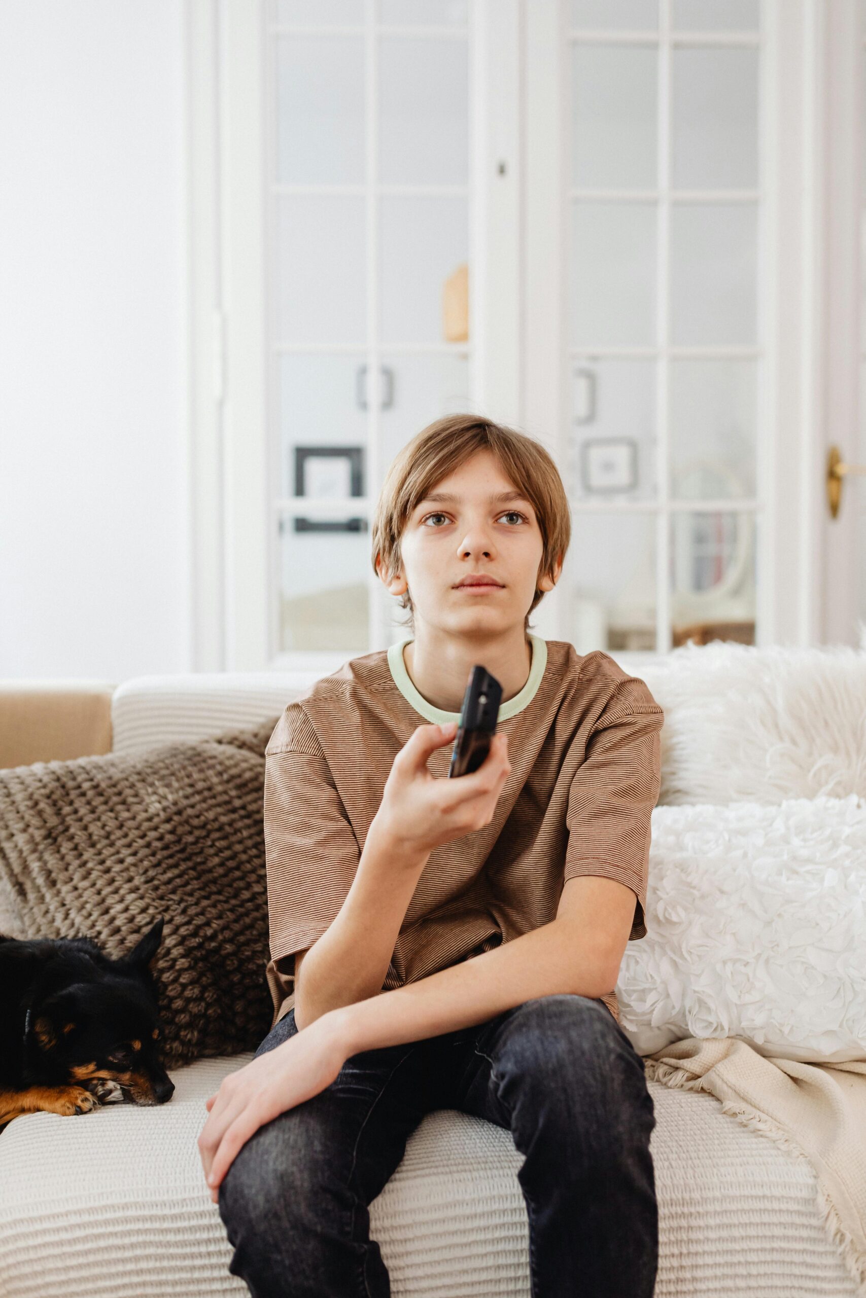 A boy holds a TV remote while sitting on a sofa with a dog beside him indoors.