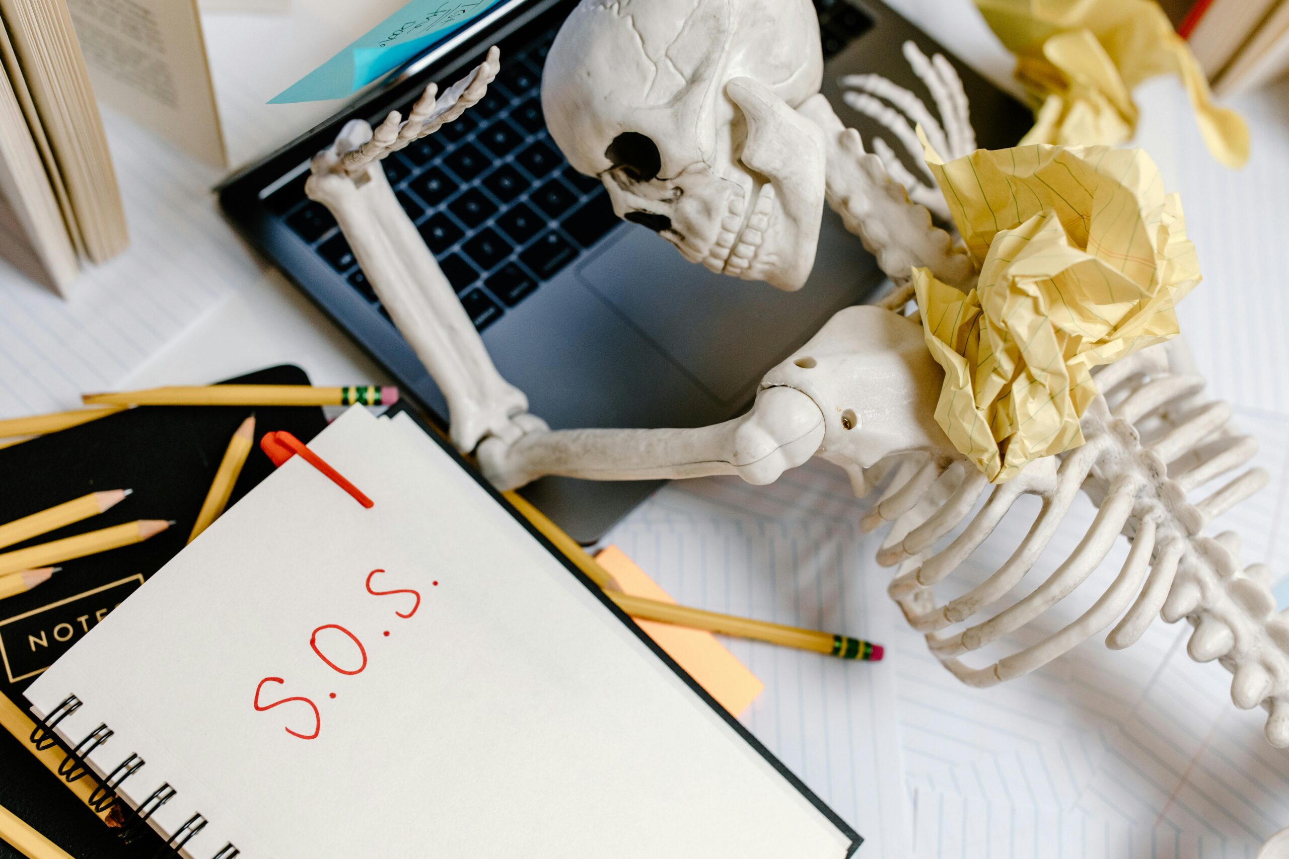 A humorous depiction of office burnout with a skeleton and S.O.S note at a cluttered desk.