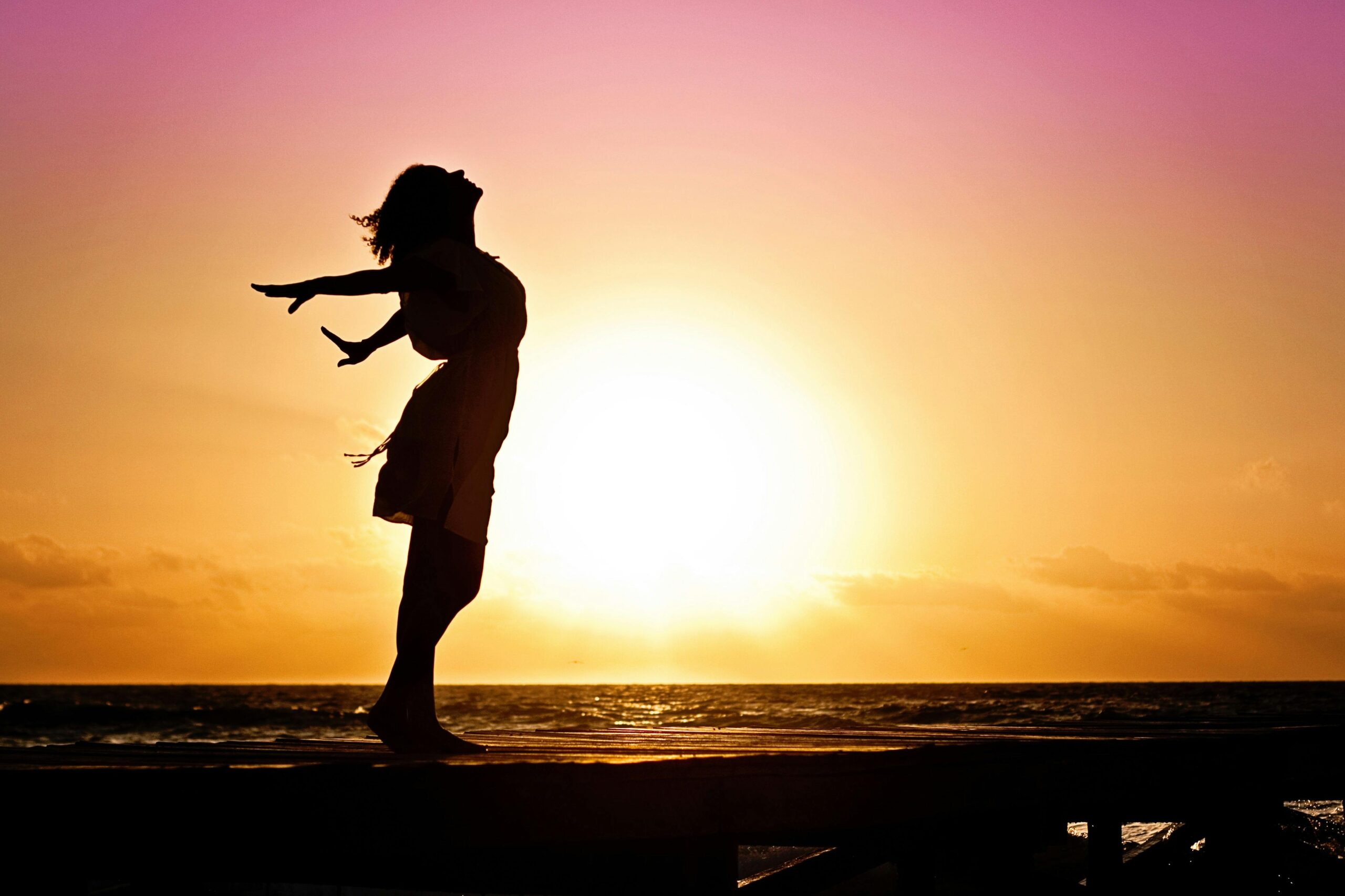 Silhouette of a woman at the beach with arms outstretched against a vibrant sunset backdrop.