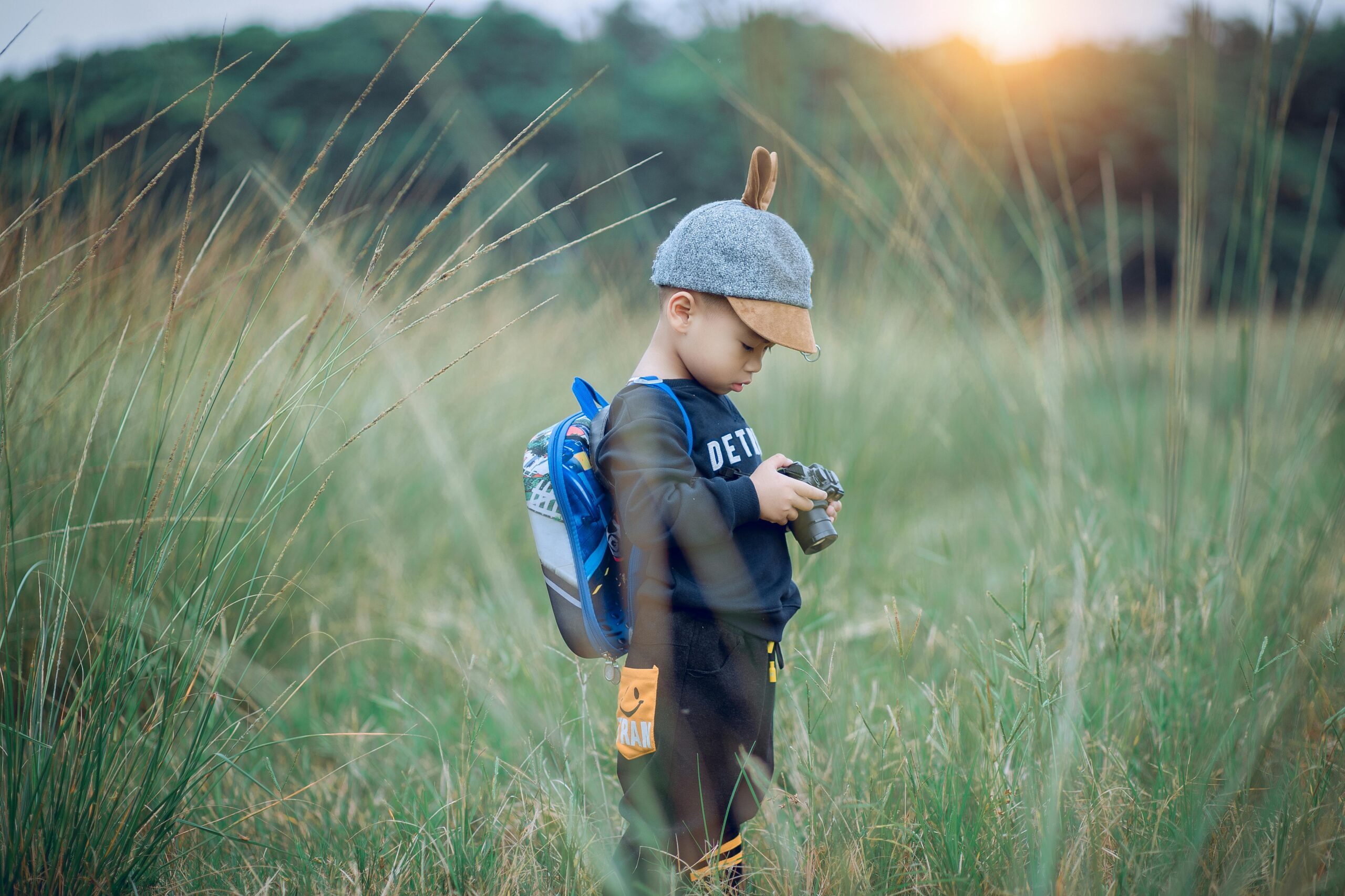 A young boy wearing a cap explores a grassy field with a camera, enjoying outdoor adventure.