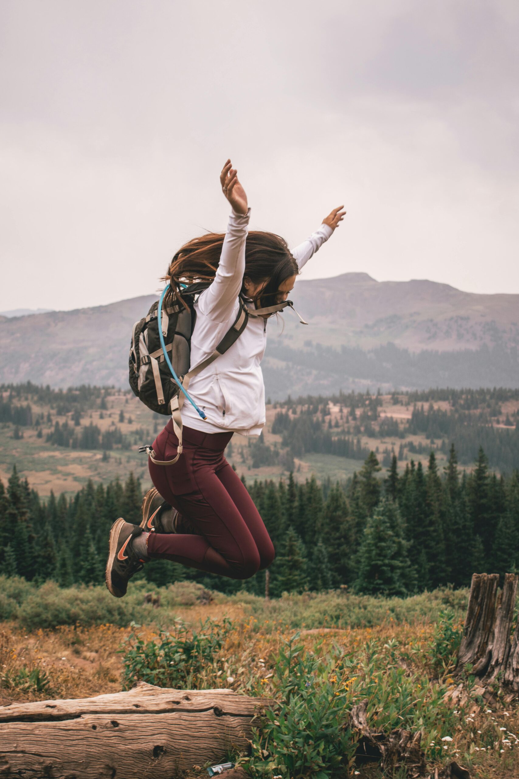 Joyful young woman leaps over a log in a scenic mountainous forest setting, embodying outdoor adventure.