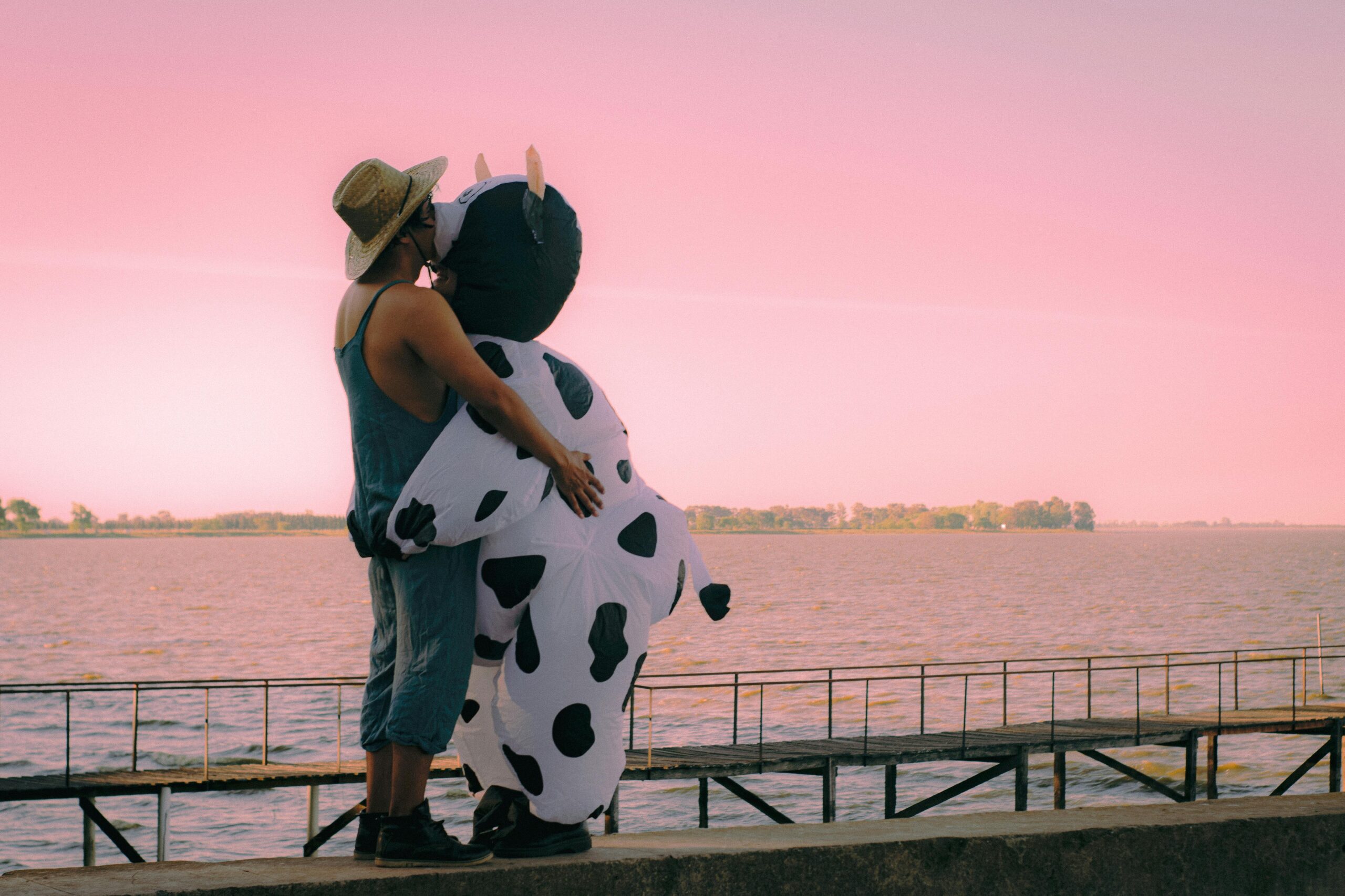 A couple shares a loving embrace at sunset, with one wearing a fun cow costume by a serene riverside.