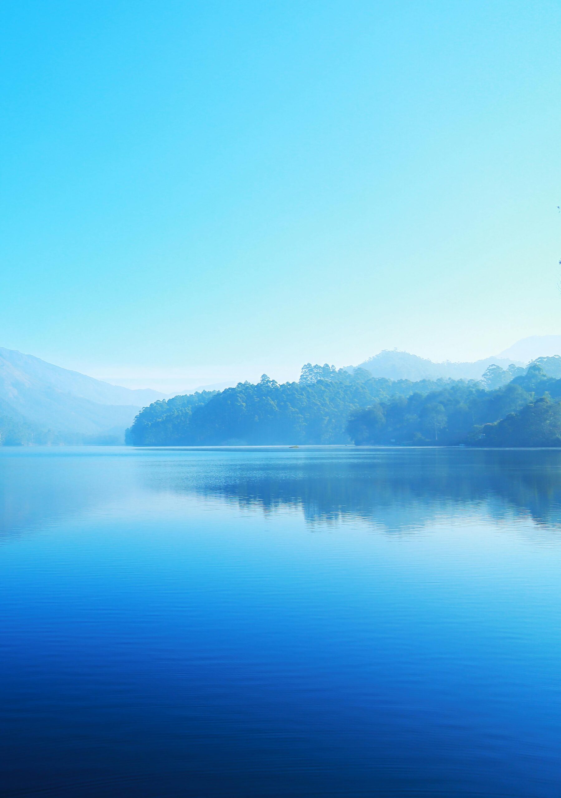 A tranquil lake reflecting misty mountains under a bright blue sky, offering a sense of calm and serenity.