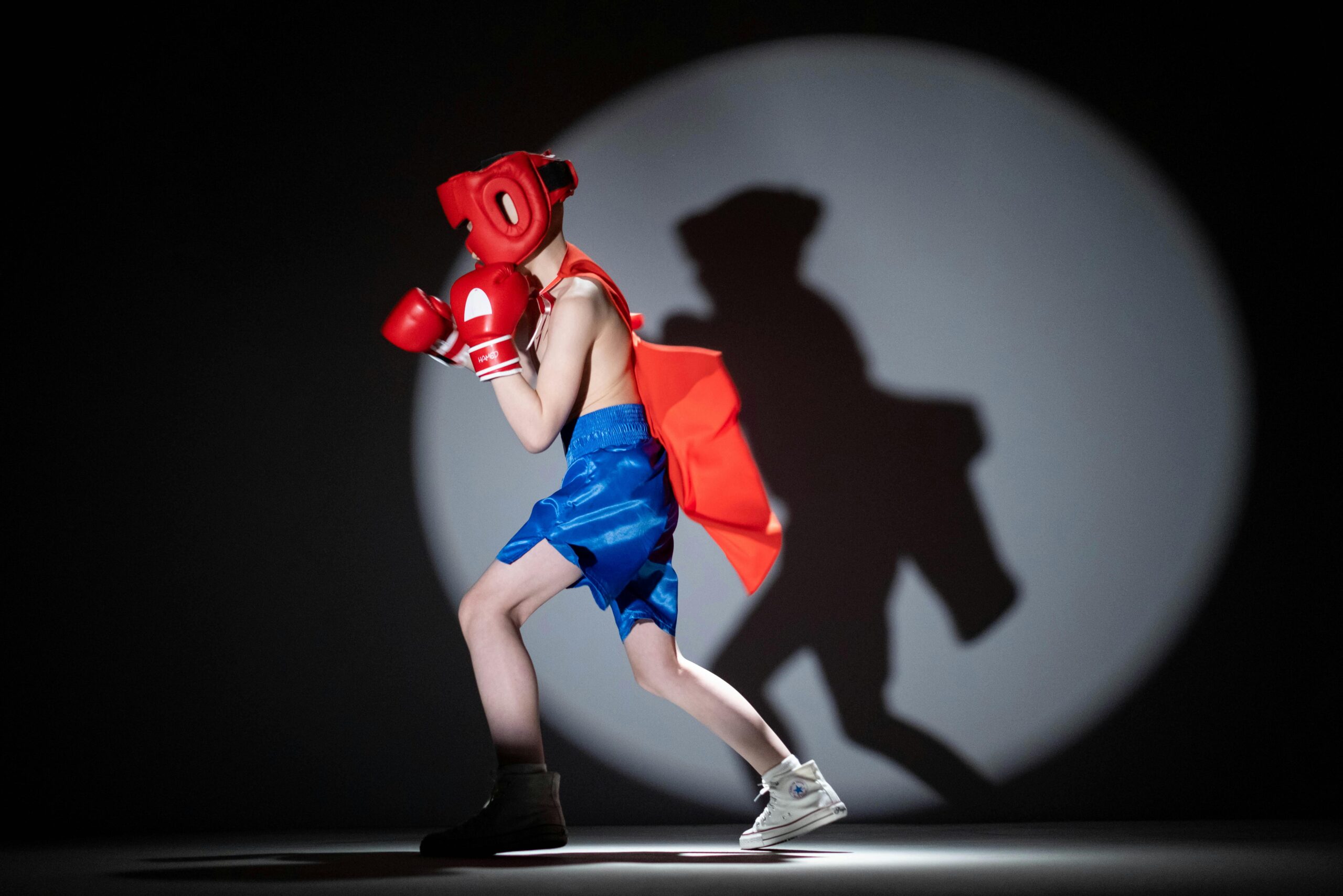 Child dressed as a superhero boxer shadow boxing under studio spotlight.