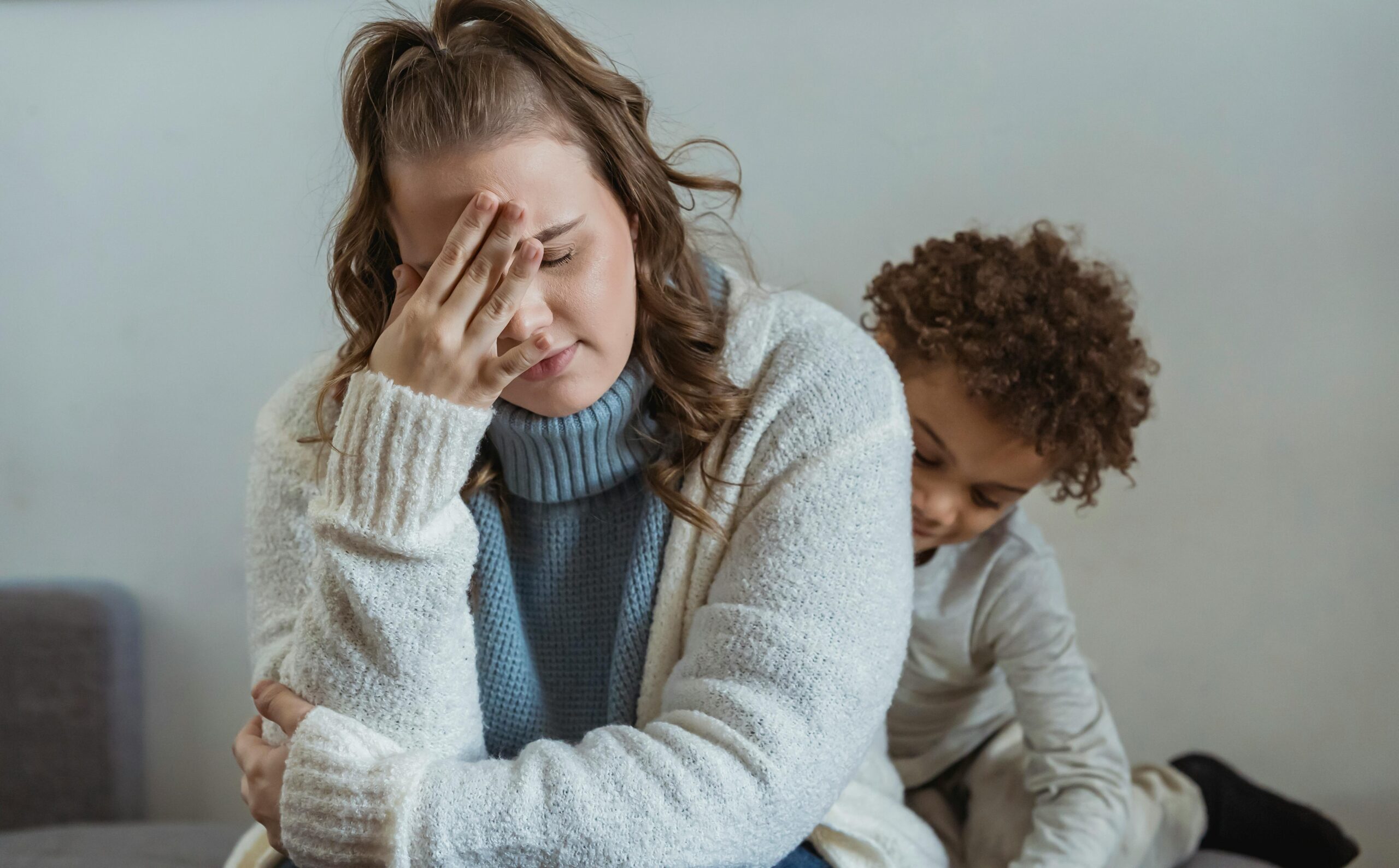 Frustrated mother with hand on forehead and closed eyes sitting near African American son near wall in room at home