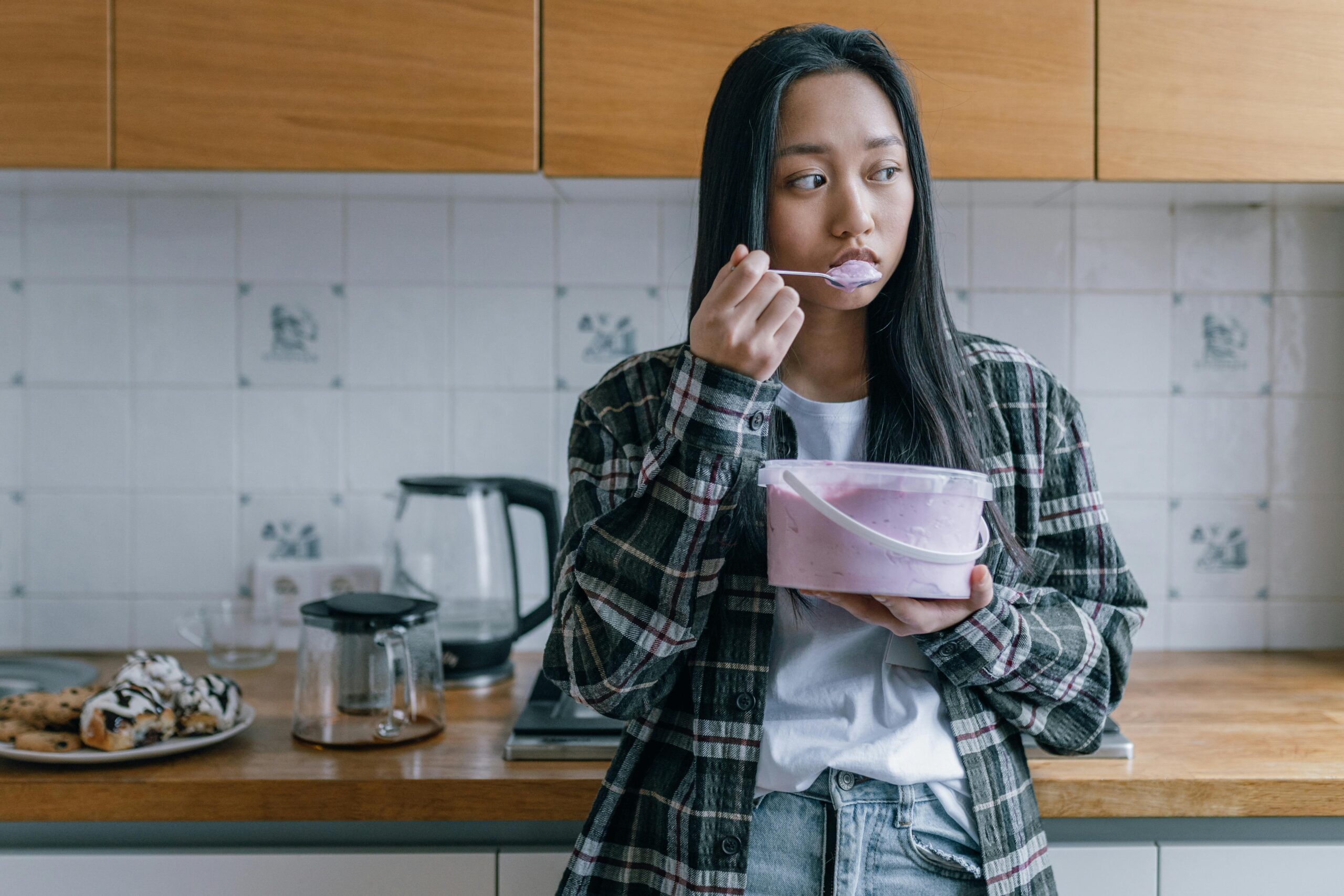 A young woman in a flannel shirt enjoys ice cream while standing in a kitchen.