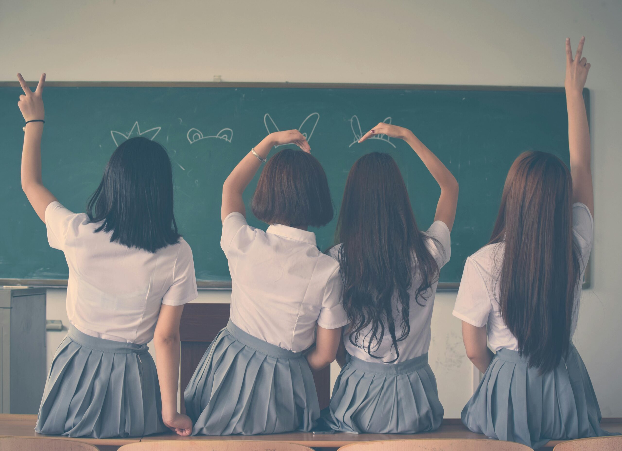 Group of teenage girls in school uniforms enjoying time together in a classroom, posing with playful gestures.