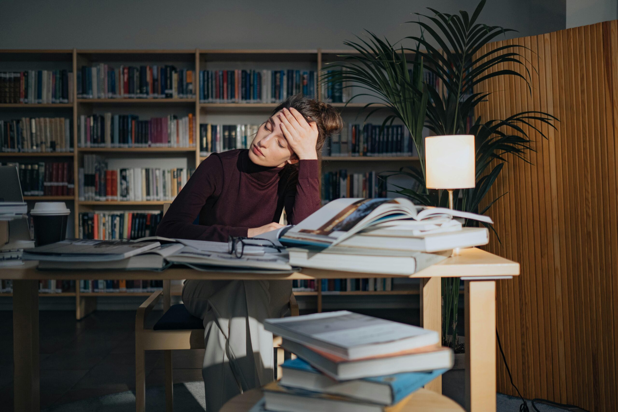 A tired woman surrounded by books, studying in a library, feeling overworked and stressed.