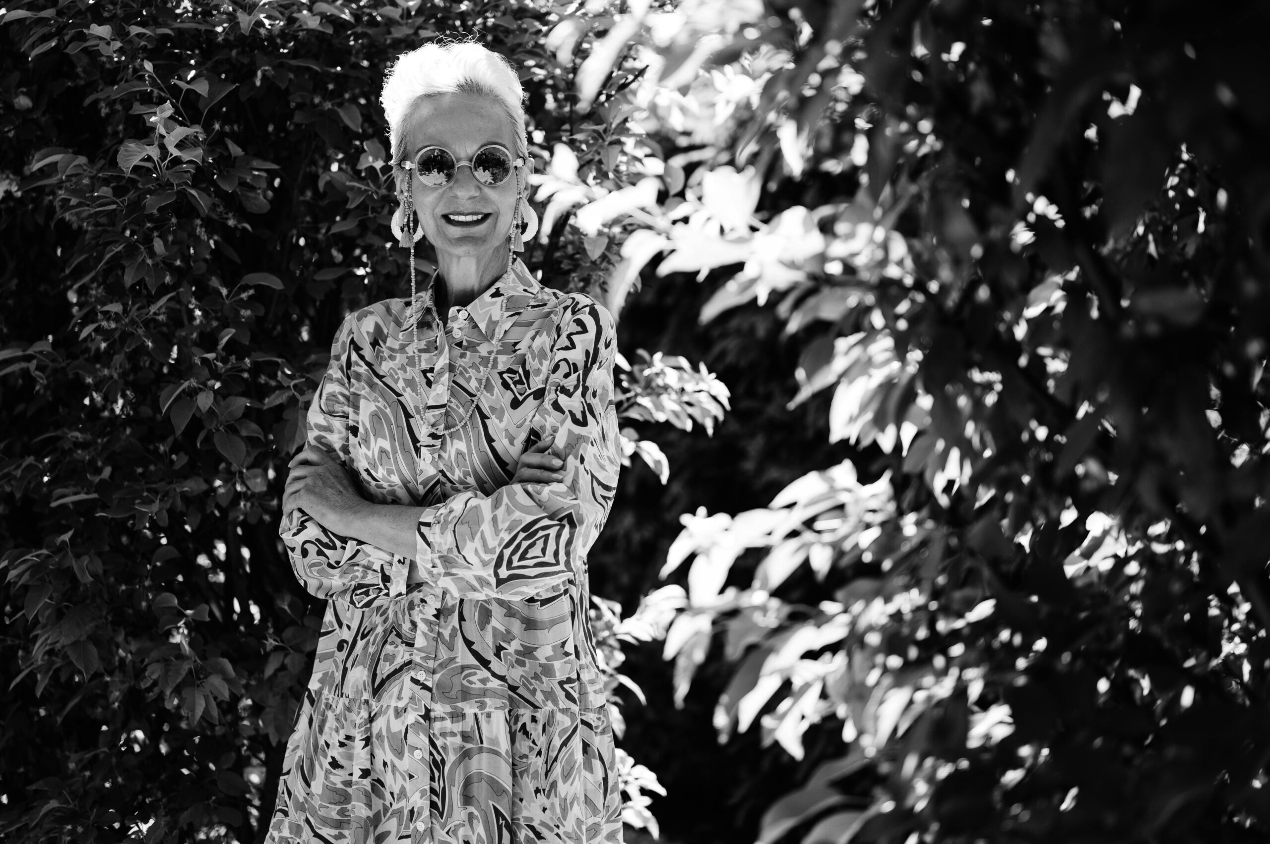 A stylish senior woman with sunglasses poses confidently outdoors in a monochrome setting.