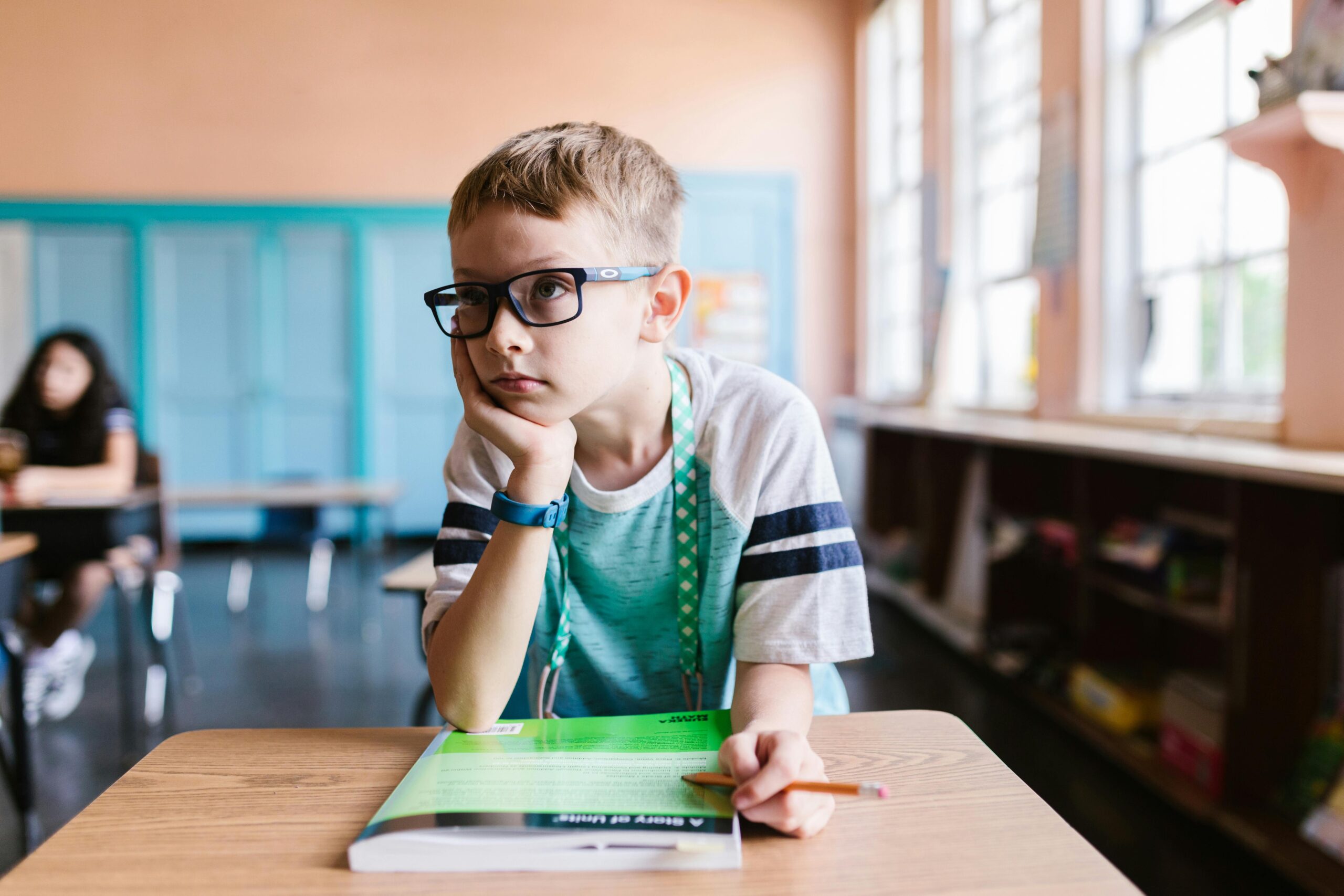 A young boy wearing eyeglasses sits at a desk in a classroom, focused on a book.